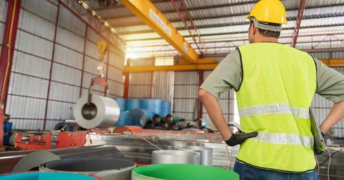 An industrial worker watching an overhead crane move a large metal coil. He has both hands on his hips.