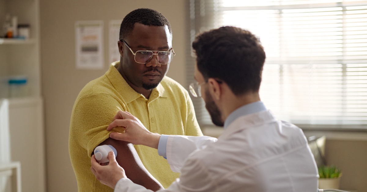 A man in a white lab coat is placing a blood sugar sensor on the arm of another man in a yellow polo in an office.