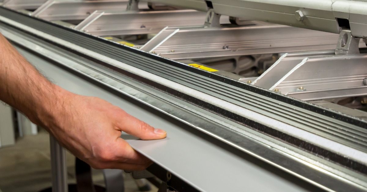 A close-up of a man's hands holding the edge of a sheet of thin metal as it enters a manual bending machine.