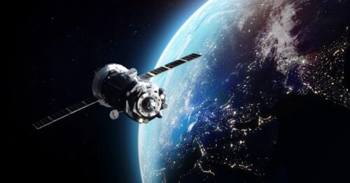 A white and black space satellite with solar panels orbiting the Earth, with half of the Earth day and the other half night.