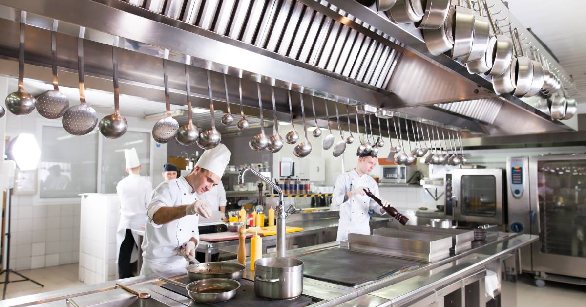 A group of chefs works in a commercial kitchen. One chef sprinkles seasoning on a pan, while another uses a large pepper shaker.