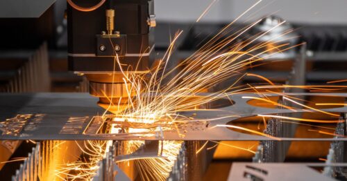 A laser cutting machine cuts a thin metal sheet in a manufacturing plant. Sparks fly while the machine cuts an intricate pattern.