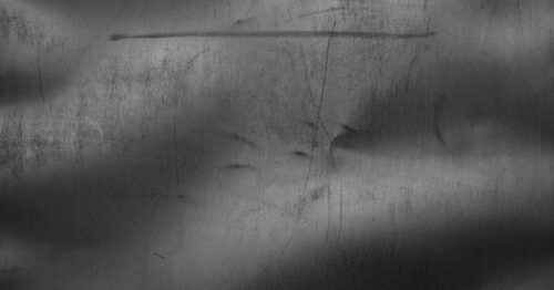 A close up view of some sheet metal. The metal surface has a few scratches across it and a dent in the middle.