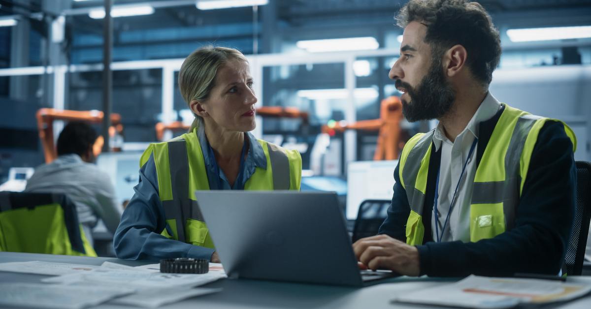 A man and woman talking to each other while sitting at a desk with a laptop. They are both wearing reflective vests.
