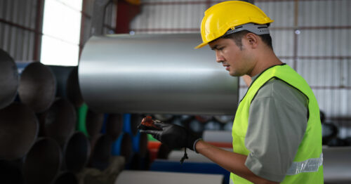 An industrial worker standing in front of some stacks of sheet metal coils with a device in his hand.