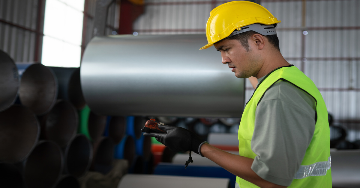 An industrial worker standing in front of some stacks of sheet metal coils with a device in his hand.