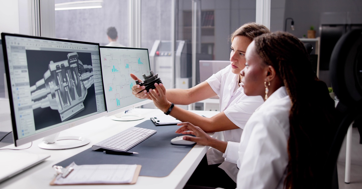 Two women looking at a couple of monitors that show the mechanisms of a metal part. One of them is holding the actual part.