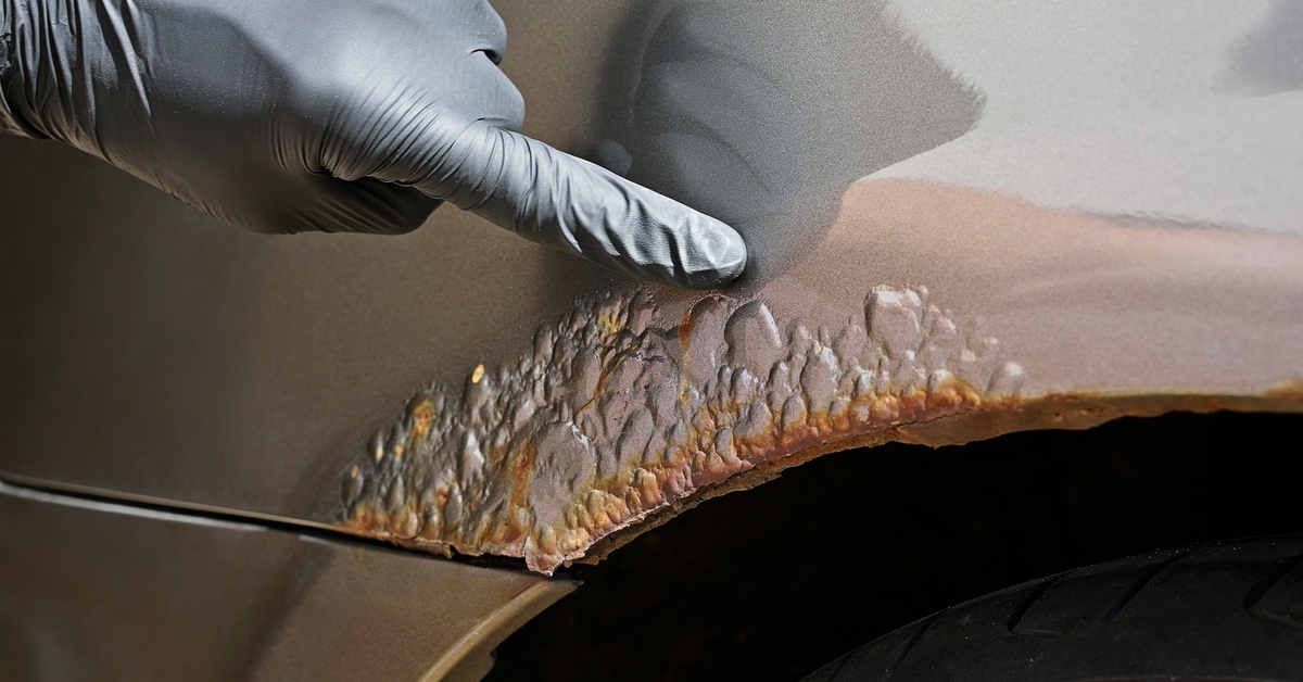 A gloved finger pointing at some rust on the side of a car. The metal that's rusting is right over the wheel.