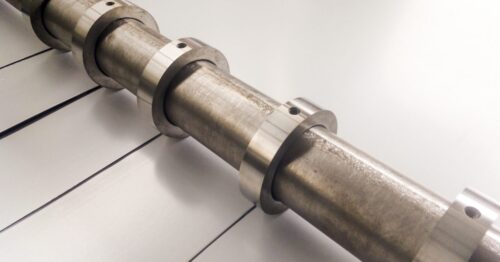 A close-up view of a metal roller cutter slicing a shiny sheet into narrow strips as it moves through the machine.